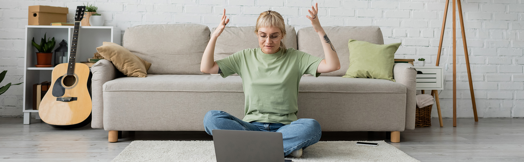 Blonde tättowierte Frau im Schneidersitz am Boden mit Laptop aufgeregt emotional, modernes Wohnzimmer, Couch, Teppich